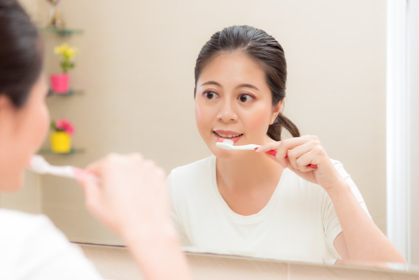 young smiling housewife using toothbrush cleaning teeth after eating food or waking up in morning standing on bathroom looking mirror brushing.